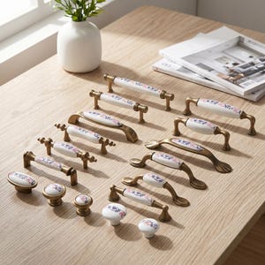 May include: A collection of cabinet hardware including various drawer pulls and knobs. Each piece features a white ceramic center with a delicate blue and pink floral pattern, accented by antique brass-toned metal ends and mounting bases.