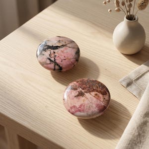 May include: Two polished, circular stones with smooth surfaces. The top stone features a blend of soft pink hues with prominent black veining. The bottom stone displays a mix of deep reddish-brown and pink tones with subtle, fine white mineral lines.