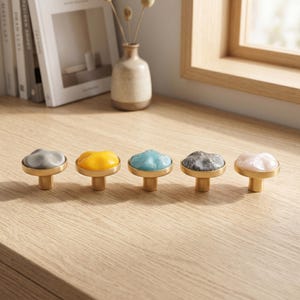 May include: Five cabinet knobs with gold-toned metal bases and rounded, irregular tops. The tops feature distinct finishes: solid matte gray, glossy yellow, glossy light blue, gray marble-like pattern, and a pale, translucent off-white.
