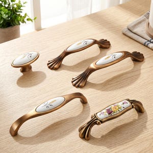 May include: A collection of five cabinet hardware pieces including one oval knob and four arched handles. The hardware features antique brass-toned metal bases with white ceramic inserts decorated with floral and wheat-like botanical motifs.