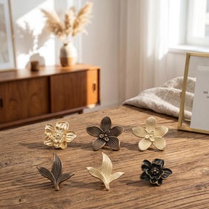 Può includere: Sei pomelli decorativi per mobili a forma di fiore, in finiture oro, bronzo e nero, sono esposti su una superficie in legno. I pomelli presentano dettagliati motivi floreali e fogliari. Ideali per l'arredamento della casa.