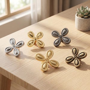 May include: Five metal cabinet knobs shaped like four-petaled flowers. The set includes one polished silver, one bright gold, one dark charcoal, one matte gold, and one antique bronze finish. Each knob features a central square and four petals.
