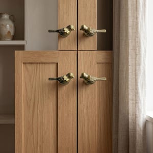 May include: Four brass-colored metal cabinet knobs shaped like small birds with detailed wing textures. These bird-shaped pulls are mounted on light-toned wooden cabinet doors, featuring a metallic finish and a horizontal orientation for each piece.