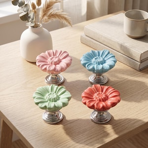 Op de afbeelding: Een set van vier keramische ladeknoppen in de vorm van bloeiende bloemen in pastelroze, lichtblauw, mintgroen en koraaloranje. Elke knop heeft een glanzende afwerking en is gemonteerd op een ronde, gepolijste zilverkleurige metalen voet.