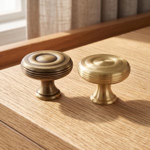 Puede incluir: Dos pomos redondos para armarios con un diseño circular escalonado. Un pomo presenta un acabado de latón antiguo oscuro con pátina visible, mientras que el otro tiene un acabado dorado cepillado brillante. Ambos pomos tienen una base acampanada.