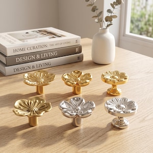 Op de afbeelding: Een verzameling van zes metalen kastknoppen in de vorm van bloemen, gerangschikt in twee rijen. Drie knoppen hebben een gepolijste gouden afwerking, terwijl de andere drie een reflecterende zilveren afwerking hebben. Elke knop heeft een steel.