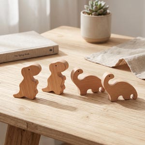 Puede incluir: Cuatro figuras de dinosaurios de madera de color claro dispuestas en fila. Dos figuras tienen forma de terópodos bípedos con brazos cortos y dos tienen forma de saurópodos de cuello largo. Cada pieza tiene un acabado liso de veta de madera.