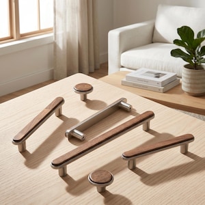 May include: A collection of modern cabinet hardware, including knobs and pulls. The hardware features a combination of brushed metal and dark wood. The knobs are round, and the pulls are rectangular. The hardware is displayed on a light wood surface.