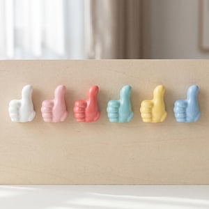 May include: A set of six ceramic thumb-up shaped hooks in a row, featuring a smooth glossy finish in white, light pink, coral, turquoise, yellow, and light blue colors, each designed with a curved thumb pointing upward for hanging small items.