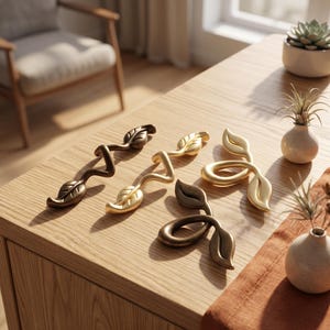May include: Four metal cabinet pulls shaped like branches with leaves. Two pulls are long and wavy with a dark bronze and a gold finish. Two pulls are shorter and looped with a dark bronze and a gold finish, all featuring detailed leaf accents.