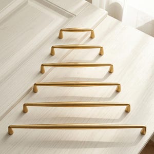 May include: A set of six gold-toned metal cabinet pulls arranged in a pyramid formation, each featuring a smooth, elongated, slightly curved bar design with rounded ends and a brushed finish, displayed in increasing lengths from top to bottom.