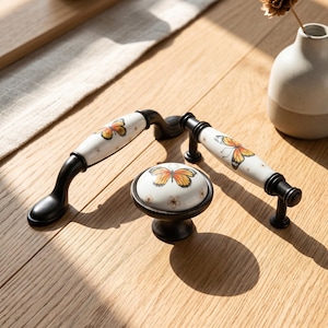 May include: Two arched drawer pulls and one round cabinet knob made of white ceramic with black metal bases. Each piece features a painted orange and black butterfly design with small white floral accents on the white ceramic surface.