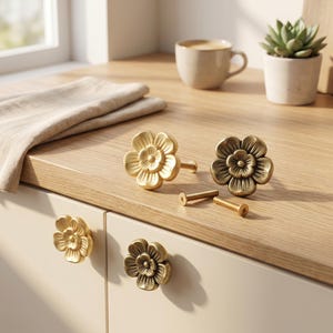 Può includere: Quattro pomelli per armadietti color ottone a forma di fiori a cinque petali. Due pomelli sono fissati su ante di armadietti chiare, mentre altri due pomelli e due viti in metallo sono appoggiati sul piano di lavoro in legno.