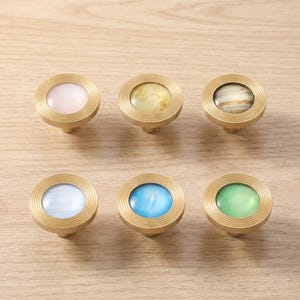 May include: A set of six circular cabinet knobs featuring a brushed gold metal frame with concentric ridged detailing. Each knob contains a smooth, rounded center inlay in distinct colors: pale pink, marbled yellow, brown, light blue, bright blue, and green.