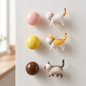 May include: A set of three magnetic hooks featuring small cat figurines and three smooth, round spheres in pink, yellow, and brown. Each cat has a collar and a raised tail, while the spheres are glossy and solid in color, arranged vertically.
