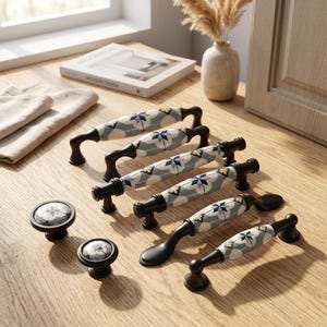 May include: A collection of cabinet hardware including four arched drawer pulls and two round knobs. Each piece features a ceramic center with a gray, white, and yellow geometric star pattern, contrasted by matte black metal ends and mounting bases.