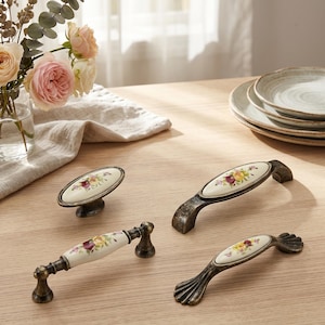 Könnte beinhalten: Vier Möbelgriffe und Knöpfe mit einem ovalen Keramikzentrum, das ein florales Muster aus gelben, rosa und roten Rosen zeigt. Die Beschläge haben ein antikes Bronze-Finish mit dekorativen Metalldetails an den Enden und Befestigungspunkten.