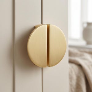 Può includere: Un pomello per mobile circolare con finitura in oro spazzolato, caratterizzato da una fessura verticale al centro che divide la forma rotonda in due metà uguali. La ferramenta è montata su un'anta di un mobile a pannelli di colore chiaro.