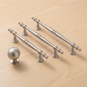 Può includere: Un set di tre maniglie per armadietti in nichel spazzolato con un design affusolato ad anelli multipli e un pomello sferico abbinato. Ogni maniglia presenta due basi di montaggio circolari, mentre il pomello ha una base piatta e rotonda.
