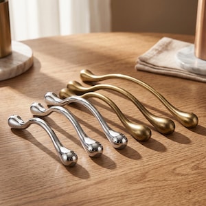 May include: A collection of decorative cabinet pulls in gold and silver tones. The curved handles have a teardrop-shaped end. The hardware is arranged on a wooden surface, showcasing their design and finish.