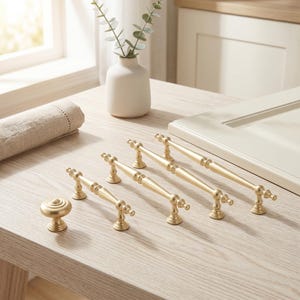May include: A collection of gold-toned cabinet hardware including one round knob and three elongated drawer pulls. Each piece features a smooth, metallic finish with a central cylindrical bar and decorative tapered ends supported by two small pedestals.