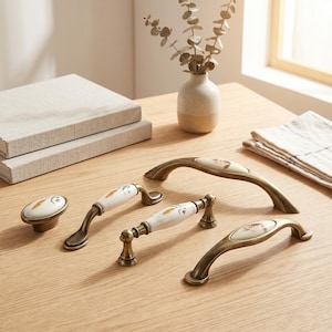 May include: A collection of four cabinet hardware pieces featuring antique brass metal finishes and white ceramic inserts with delicate floral patterns. The set includes one oval knob, two arched handles, and one straight handle with decorative ends.