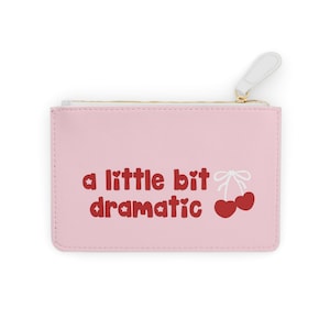 May include: A pink zippered pouch with the phrase "a little bit dramatic" in red script. The design includes red cherries with white bows. The zipper pull is white with a gold-colored zipper.