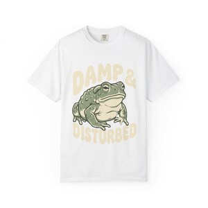 Puede incluir: Camiseta blanca con un dibujo de rana verde y gris y el texto "DAMP & DISTURBED" en amarillo claro. La rana está centrada en la camiseta, con el texto arriba y abajo.