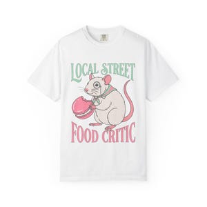 May include: White t-shirt featuring a cartoon rat wearing a monocle and holding a pink macaron. The shirt has the text "LOCAL STREET FOOD CRITIC" in green and pink lettering.