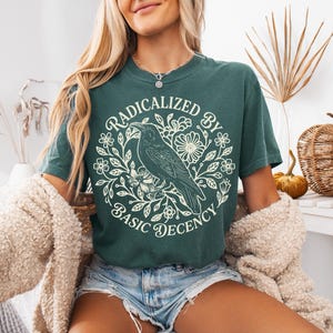 Op de afbeelding: Donkergroen t-shirt met een witte afbeelding van een raaf op een bloemtak, omringd door de tekst "RADICALIZED BY BASIC DECENCY". Het shirt heeft een relaxte pasvorm.