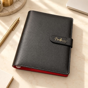 May include: A black leather-textured planner with a secure strap closure featuring the gold embossed text Nexella Luxe. The planner has a vibrant red interior lining visible along the bottom edge and is accompanied by a gold-toned metal pen.