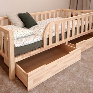 Pode incluir: Uma estrutura de cama de madeira clara com uma grade de segurança de ripas em três lados e duas grandes gavetas de armazenamento deslizantes por baixo. A cama está coberta com um lençol branco estampado, uma almofada verde-escura e uma cinzenta-clara.
