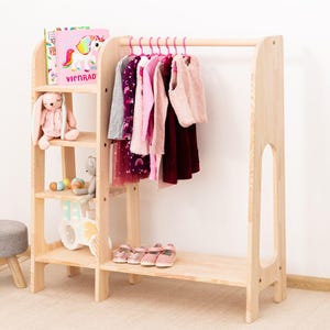 Peut inclure: Porte-vêtements pour enfants en bois clair avec étagères et bibliothèque intégrée. Des cintres roses soutiennent des vêtements. Un lapin rose en peluche et des livres sont sur les étagères. Un petit tabouret gris et des chaussures roses sont également visibles.