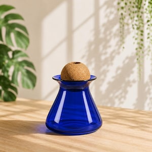 May include: A cobalt blue glass vase with a cork stopper. The vase has a flared rim and a rounded base. The cork stopper is a light brown color and has a small hole in the center. The vase is sitting on a wooden surface.