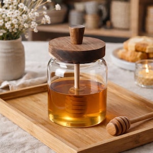 Peut inclure: Un pot à miel en verre avec un couvercle et un bâtonnet en bois, rempli de miel doré. Le pot est posé sur un plateau en bois, avec un autre bâtonnet à miel à côté. Un petit vase de fleurs blanches est en arrière-plan.