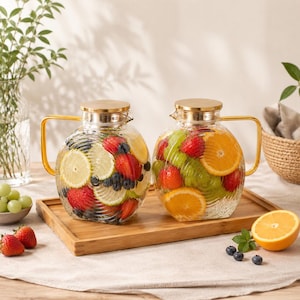 May include: Two glass pitchers filled with sliced fruit, including strawberries, oranges, and lemons. Each pitcher has a gold handle and lid. They sit on a wooden tray, with additional fruit and greenery nearby.