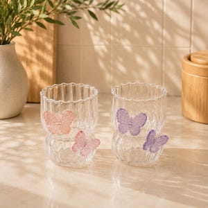 Peut inclure: Deux verres transparents avec une texture côtelée verticale et un milieu arrondi et resserré. Chaque verre présente deux décorations papillon en relief et texturées fixées à l'extérieur, l'une dans un ton pêche doux et l'autre violet.