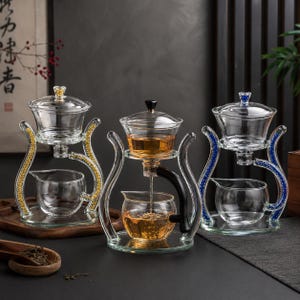 Può includere: Tre set per infusione di tè in vetro con supporti curvi. Il set a sinistra ha dettagli dorati, quello centrale ha un manico nero e vetro trasparente, e quello a destra ha dettagli blu. Ogni set include una camera di infusione superiore e una caraffa.