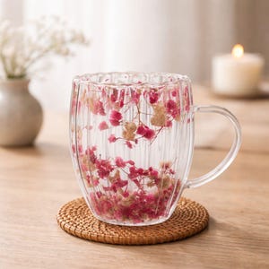 Peut inclure: Une tasse en verre transparent avec une poignée, remplie de fleurs séchées roses et beiges. La tasse a un design côtelé et repose sur un dessous de verre rond et tissé. Un petit vase avec des fleurs blanches et une bougie allumée sont en arrière-plan.