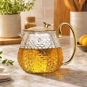 May include: A clear glass teapot with a textured scale pattern on the body, featuring a gold-coloured metal lid with a small handle and a matching curved gold-coloured metal handle. The teapot is filled with a golden-yellow liquid tea.