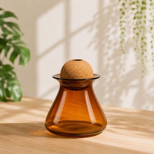 May include: Amber glass vase with a cork sphere stopper. The vase has a wide base that tapers to a narrow neck and flares out at the top. The cork stopper is a light brown color and fits snugly in the vase's opening. The vase is sitting on a wooden surface.