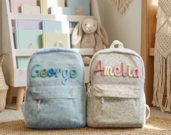 Op de afbeelding: Twee gepersonaliseerde rugzakken. Eén is lichtblauw met de naam "George" in kleurrijke letters, de andere gebroken wit met "Amelia" in roze en geel. Beide hebben een voorvak en witte bandjes, op een geweven vloerkleed.