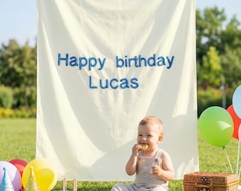 Op de afbeelding: Een kind eet een koekje voor een spandoek met de tekst "Happy birthday Lucas" in blauwe letters. Kleurrijke ballonnen, feesthoedjes en een rieten mand staan op het gras.