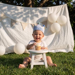 Op de afbeelding: Een baby met een lichtblauwe kroon met de naam "Ethan" in blauw geborduurd, zit op een kleine witte kruk. De baby glimlacht en houdt een cupcake vast. Witte ballonnen en een banner zijn op de achtergrond, op een groen gazon.