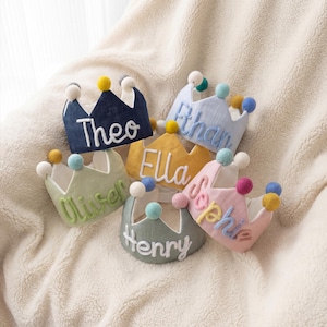 Op de afbeelding: Een verzameling kleurrijke stoffen kronen, elk geborduurd met een naam: Theo, Ethan, Ella, Oliver, Henry en Sophie. De kronen zijn versierd met pompons en liggen op een zacht, cr&egrave;mekleurig oppervlak.