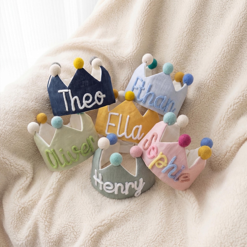 Op de afbeelding: Een verzameling kleurrijke stoffen kronen, elk geborduurd met een naam: Theo, Ethan, Ella, Oliver, Henry en Sophie. De kronen zijn versierd met pompons en liggen op een zacht, cr&egrave;mekleurig oppervlak.
