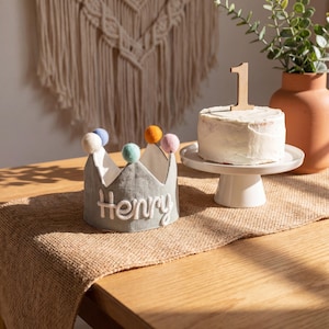 Op de afbeelding: Een grijze stoffen kroon met de naam "Henry" in het wit geborduurd, bekroond met kleurrijke pompons. Een kleine taart met een houten nummer "1" topper staat op een witte taartstandaard. De sc&egrave;ne speelt zich af op een houten tafel met een jute loper.