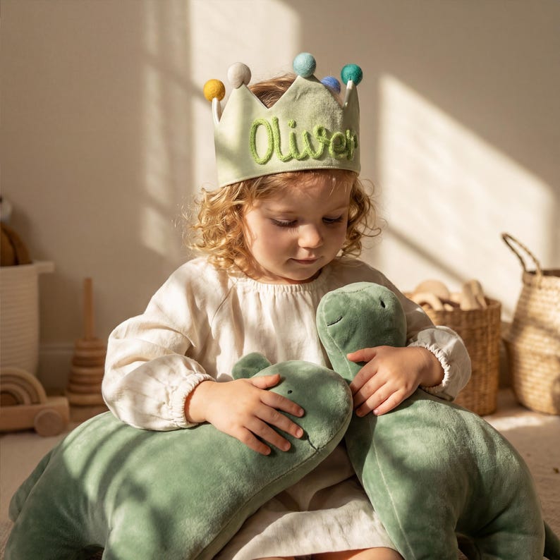 Op de afbeelding: Een kind draagt een lichtgroene kroon met de naam "Oliver" in groen geborduurd. De kroon is versierd met kleurrijke pompons. Het kind houdt twee groene dinosaurusknuffels vast.
