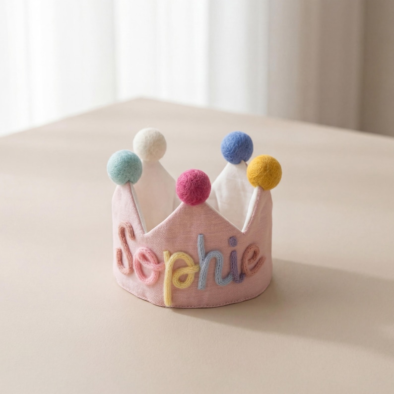 May include: A pink fabric crown with the name "Sophie" embroidered in colorful thread. The crown is topped with six colorful pom-poms. The crown is sitting on a light-colored surface.