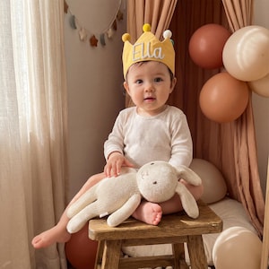 Op de afbeelding: Een baby met een gele kroon met de naam "Ella" erop geborduurd. De baby zit op een houten kruk en houdt een knuffelkonijn vast. Ballonnen in bruine en cr&egrave;metinten zijn op de achtergrond te zien.